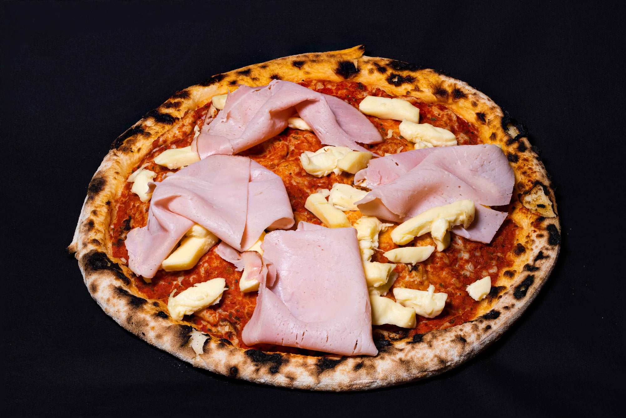 A wood-fired pizza with fresh toppings including sliced ham and cheese, presented on a dark background.