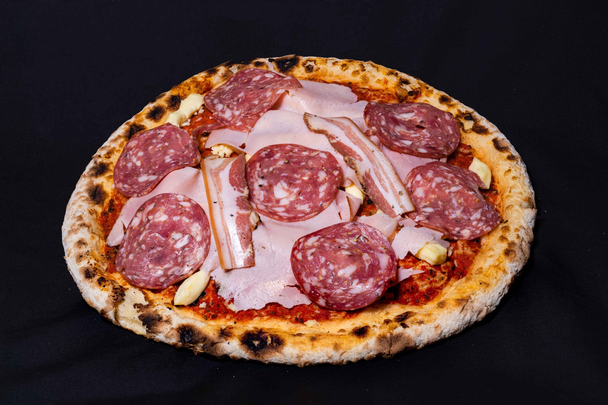 A wood-fired pizza with a meaty topping including ham, salami, and pancetta, on a dark background.