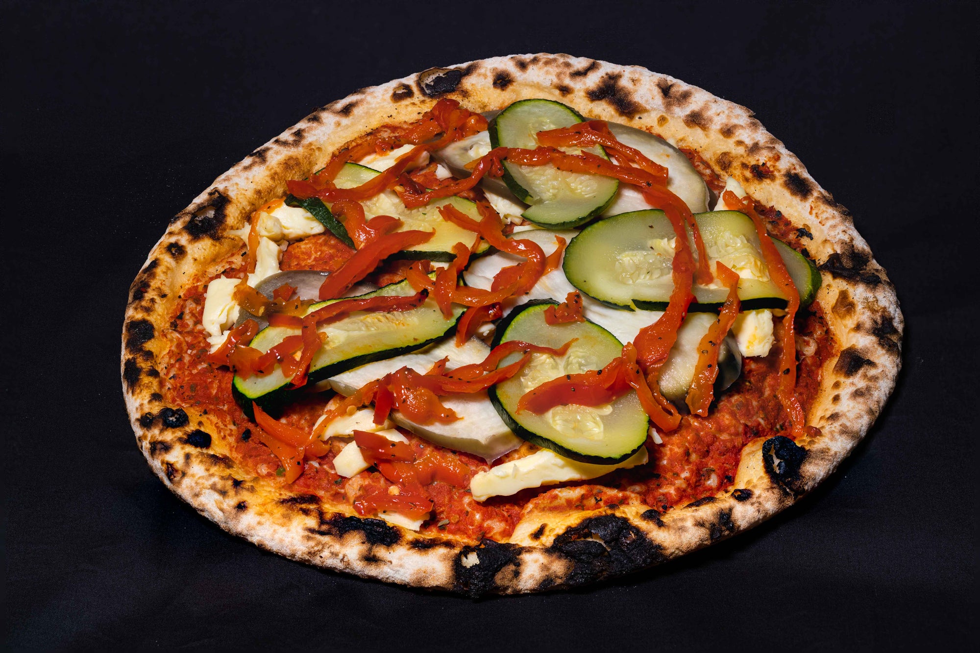A wood-fired pizza with vegetarian toppings including zucchini, eggplant, and capsicum, lying on a tomato sauce base and fresh mozzarella cheese.